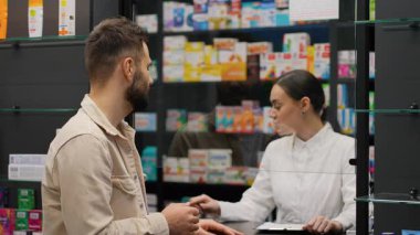 Genç adam eczanedeki kadın eczacıyla konuşuyor. Profesyonel kimyager erkek müşteriye tavsiye veriyor ve ilaç satıyor.