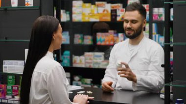 Genç bir erkek eczacı tezgahın arkasında durup bayan bir müşteriyle konuşuyor. Reçeteli bir ilacı tavsiye ediyor ve açıklıyor.