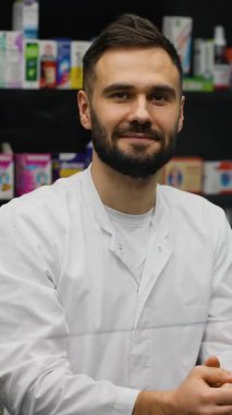 Genç, yakışıklı, sakallı, kameraya bakarken gülümseyen bir eczacı. Eczanede kendine güvenen bir sağlık uzmanı