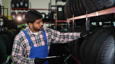 Üniformalı otomobil servisi çalışanı bir not panosuna not alıyor. Erkek çalışan stokları yönetiyor ve garajdaki lastik lastikleri inceliyor.