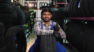 Mavi tulumlu dost canlısı Hintli tamirci yeni bir lastik öneriyor. Tatmin olmuş oto hizmet çalışanı gülümsüyor ve işaret veriyor.