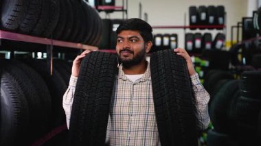 Araba tamirhanesinde iki yeni araba lastiği tutan neşeli Hintli adam. Memnun erkek müşteri aracı için en iyi seçeneği seçiyor.