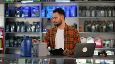 Araba parçaları dükkanında tezgahın arkasında duran erkek işçi. Genç yönetici pil ve motor yağını kontrol ediyor, panoya yazıyor ve gülümsüyor.