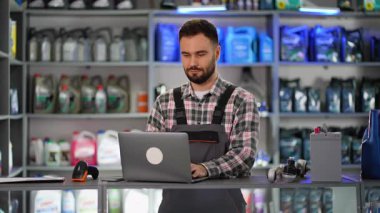 Bir oto yedek parça mağazasında envanteri kontrol etmek için dizüstü bilgisayar kullanan ve sonra kameraya gülümseyerek süspansiyon bileşeni sunan bir erkek satıcı.