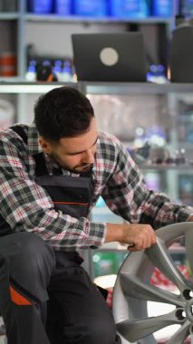 Profesyonel otomobil tamircisi dikkatli bir şekilde araba parçası dükkanında gümüş alaşımlı yeni bir tekerleği kontrol ediyor, sonra kameraya gülümsüyor.