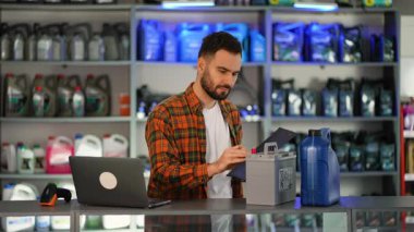 Bir oto yedek parça dükkanında çalışan genç bir erkek envanteri kontrol ediyor. Bir araba aküsünü ve bir kutu motor yağını tarıyor, sonra da not alıyor.