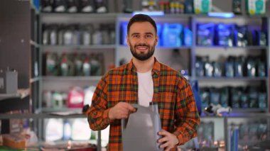 Ekose gömlekli, kendine güvenen sakallı adam kameraya gülümsüyor. Bir araba parçası mağazasında gri bir motor yağı kutusu sunuyor.