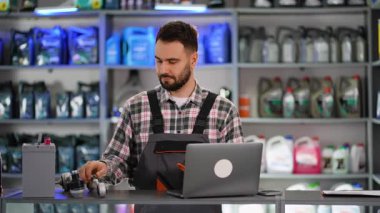 Araba parçası mağazasında çalışan üniformalı profesyonel bir erkek satıcı. Çalışan otomotiv parçalarını tarıyor ve bir dizüstü bilgisayar kullanıyor.
