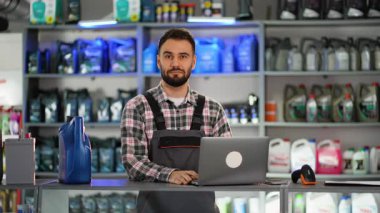 Dost canlısı erkek araba tamircisi tezgahta dizüstü bilgisayarda çalışıyor ve bir oto yedek parça mağazasında sentetik motor yağı kutusu sunuyor.
