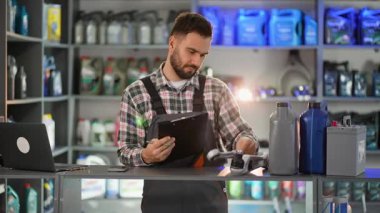 Üniformalı bir erkek araba tamircisi bir oto yedek parça mağazasında çalışıyor, envanteri kontrol ediyor ve tezgahın arkasındaki panoya not alıyor.