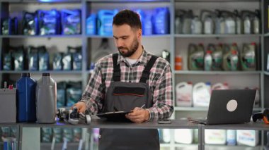 Araba yedek parça dükkanında çalışan üniformalı profesyonel araba tamircisi. Adam envanteri kontrol ediyor ve gülümseyen kameraya bakıyor.