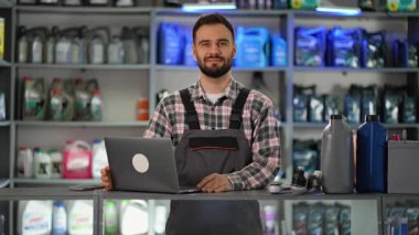 Yakışıklı erkek araba tamircisi ve üniformalı satıcı bir oto yedek parça dükkanında dizüstü bilgisayarla çalışıyor, sonra da kameraya gülümsüyor.
