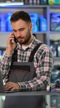 Üniformalı profesyonel otomobil tamircisi bir mağazadaki dizüstü bilgisayarda yedek parça ararken müşteriyle telefonda konuşuyor.