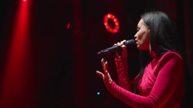 Canlı müzik performansı sırasında mikrofona şarkı söyleyen yetenekli bir kadın vokalist. Sahne ışıkları, karanlık bir kulüpteki enerjik gösterisini aydınlatıyor.