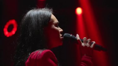 Mikrofonlu tutkulu bir bayan şarkıcı sahnede şarkı söylüyor. Kırmızı ışıklar dramatik ve duygusal bir atmosfer yaratıyor.