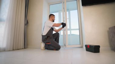 Üniformalı genç bir erkek işçi beyaz plastik bir balkon kapısının sapını tornavida kullanarak kurarken ya da tamir ederken diz çöküyor.