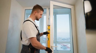 Yeni takılmış plastik pencere çerçevesinin dikey hizasını kontrol etmek için ruh seviyesini kullanan işçi tulumları içinde genç bir erkek.
