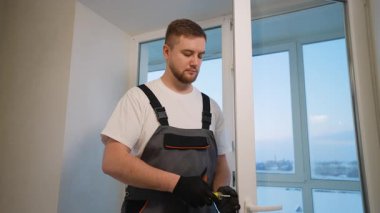 Üniformalı genç bir erkek işçi dairede tornavida kullanarak beyaz plastik pencere çerçevesine yeni bir kapı kolu yerleştiriyor ve onarıyor.
