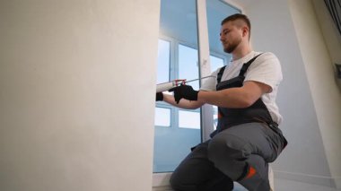 Tulumlar içinde odaklanmış genç inşaat işçisi, yeni pencere çerçevesi boyunca dikkatlice silikon dolgu maddesi uyguluyor.