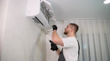 Üniformalı genç bir erkek teknisyen bir müşterinin evinde duvara monte edilmiş klima monte etmek ya da tamir etmek için elektrikli tornavida kullanıyor.