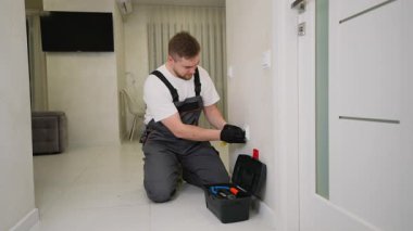 Genç elektrikçi üniformalı bir erkek modern bir daireye elektrik prizi kuruyor. El sanatları ustası alet çantasıyla elektrik kabloları üzerinde çalışıyor.