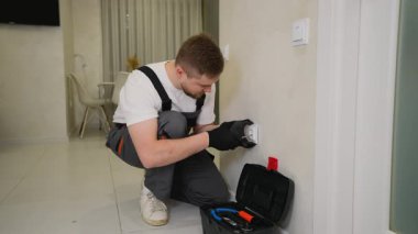 İş üniformalı genç bir elektrikçi duvar prizi kuruyor ya da tamir ediyor. Ev bakımı için araç kullanan bir tamirci.