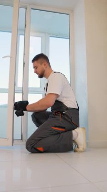 Yeni bir plastik kapı veya pencere çerçevesi kuran üniformalı genç profesyonel işçi, ev yenileme için kablosuz bir elektrikli matkap kullanıyor