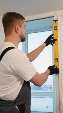 Yeni bir pencere çerçevesinin doğruluğunu kontrol etmek için ruh seviyesinde bir araç kullanan işçi tulumları içinde genç bir erkek.