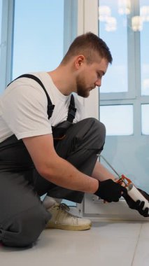 Tulum giymiş, kabartma tabancası kullanan çalışkan bir inşaat işçisi. Erkek müteahhit titizlikle silikon dolgu maddesini pencere tabanına sürüyor.