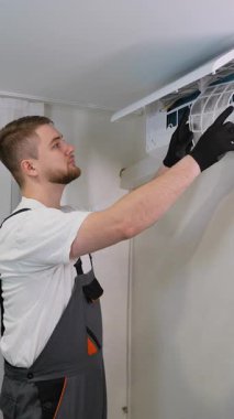 Üniforma ve eldiven giyen genç erkek teknisyen servis sırasında duvara monte edilmiş klima ünitesinin filtresini değiştiriyor veya temizliyor.