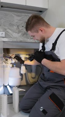 Tulumlu genç erkek teknisyen yeni bir su filtresi sistemi kuruyor. Modern bir mutfakta temiz içme suyu için profesyonel tesisat hizmeti