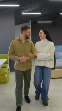 Evleri için yeni mobilyalar seçerken kol kola yürüyen mutlu genç bir çift. Modern bir galeride gülümseyen ve konuşan kadın ve erkek.