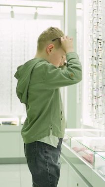 Çekici küçük çocuk modern bir optik dükkanda yeni bir gözlük seçip deniyor. Yeni görünüşüyle mutlu bir şekilde gülümsüyor.