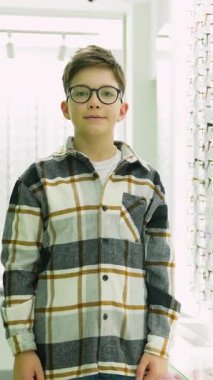 Optometristlerin kliniğinden yeni gözlük seçtikten sonra gülümseyen ve baş parmak gösteren mutlu genç çocuk.