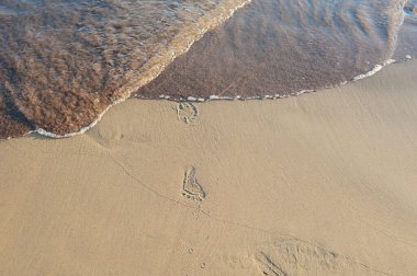Sörf, batan güneşin altında kumların üzerinde denizden gelen bebek ayak izlerini örter.
