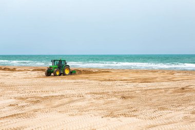 İspanya, Valencia, 29 Mart 2024: Bir traktör kumlu bir plajda çalışıyor, mavi bir gökyüzünün altında kıyı şeridi yakınında pürüzsüz yüzeyler..