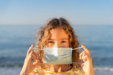 Mavi gökyüzü arka planında tıbbi maske takan mutlu bir çocuk. Kız yazın deniz kenarında eğleniyor. Coronavirus salgını ve sağlıklı yaşam tarzı konsepti