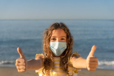 Mavi gökyüzü arka planında tıbbi maske takan mutlu bir çocuk. Kız yazın deniz kenarında eğleniyor. Coronavirus salgını ve sağlıklı yaşam tarzı konsepti