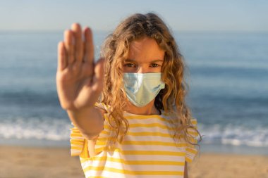 Mavi gökyüzü arka planında tıbbi maske takan mutlu bir çocuk. Kız yazın deniz kenarında eğleniyor. Coronavirus salgını ve sağlıklı yaşam tarzı konsepti