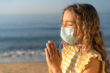 Mavi gökyüzü arka planında tıbbi maske takan üzgün bir çocuk. Sahilde dua eden bir kız. Coronavirus salgını ve sağlıklı yaşam tarzı konsepti