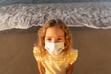 Mavi gökyüzü arka planında tıbbi maske takan üzgün bir çocuk. Kız yazın deniz kenarında eğleniyor. Coronavirus salgını ve sağlıklı yaşam tarzı konsepti