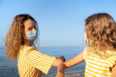 Mavi arka planda tıbbi maske takan mutlu anne ve kız. Yazın deniz kenarında eğlenen kadın ve çocuk. Coronavirus salgını ve sağlıklı yaşam tarzı aile kavramı