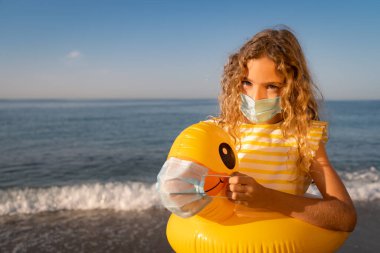 Mavi gökyüzü arka planında tıbbi maske takan mutlu bir çocuk. Yazın plajda şişme ördekli bir kız. Coronavirus salgını ve sağlıklı yaşam tarzı konsepti