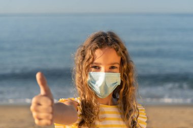Mavi gökyüzü arka planında tıbbi maske takan mutlu bir çocuk. Kız yazın deniz kenarında eğleniyor. Coronavirus salgını ve sağlıklı yaşam tarzı konsepti