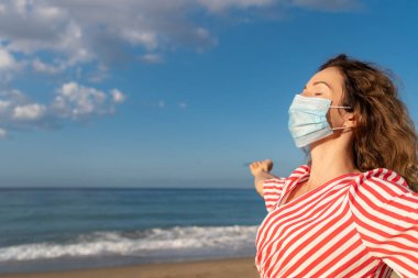Mavi gökyüzü arka planında tıbbi maske takan mutlu bir kadın. Yazın deniz kenarında eğlenen kişi. Coronavirus salgını ve sağlıklı yaşam tarzı konsepti