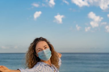 Mavi gökyüzü arka planında tıbbi maske takan mutlu bir kadın. Yazın deniz kenarında eğlenen kişi. Coronavirus salgını ve sağlıklı yaşam tarzı konsepti