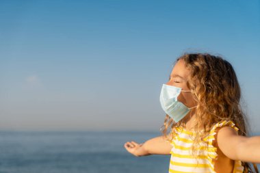 Mavi gökyüzü arka planında tıbbi maske takan mutlu bir çocuk. Kız yazın deniz kenarında eğleniyor. Coronavirus salgını ve sağlıklı yaşam tarzı konsepti