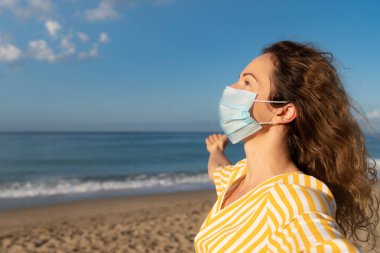 Mavi gökyüzü arka planında tıbbi maske takan mutlu bir kadın. Yazın deniz kenarında eğlenen kişi. Coronavirus salgını ve sağlıklı yaşam tarzı konsepti