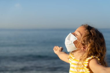 Mavi gökyüzü arka planında tıbbi maske takan mutlu bir çocuk. Kız yazın deniz kenarında eğleniyor. Coronavirus salgını ve sağlıklı yaşam tarzı konsepti