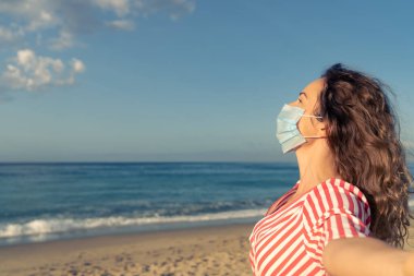 Mavi gökyüzü arka planında tıbbi maske takan mutlu bir kadın. Yazın deniz kenarında eğlenen kişi. Coronavirus salgını ve sağlıklı yaşam tarzı konsepti